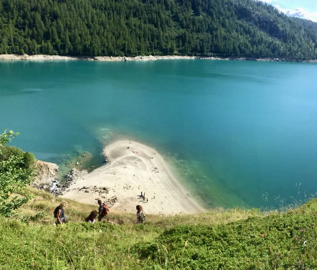 Escursionisti vicino alla spiaggia sabbiosa del Lago Ritom, con acque turchesi e bosco sullo sfondo